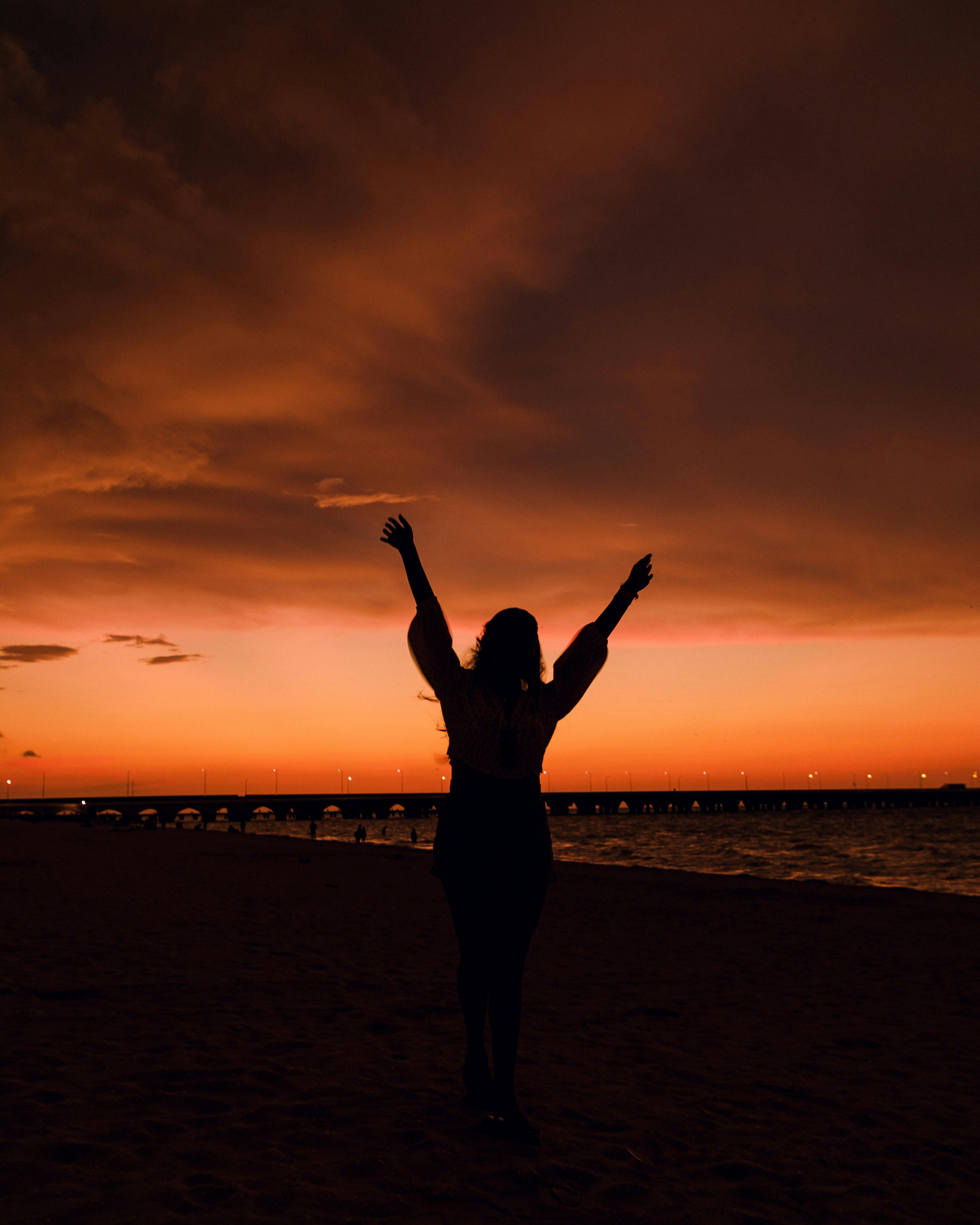 Silhouette of a person on a beach at sunset, arms raised against an orange sky.